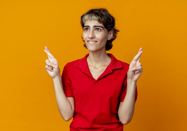 Wishing young caucasian girl with pixie haircut doing crossed fingers gesture looking straight isolated on orange background with copy space