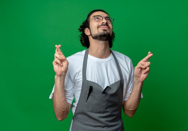 Wishing young caucasian male barber wearing glasses and wavy hair band in uniform looking up and crossing fingers isolated on green background with copy space