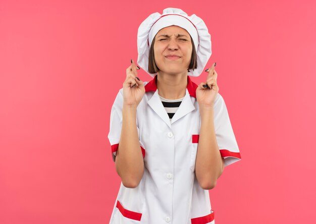 Wishing young female cook in chef uniform with crossed fingers and closed eyes isolated on pink background with copy space