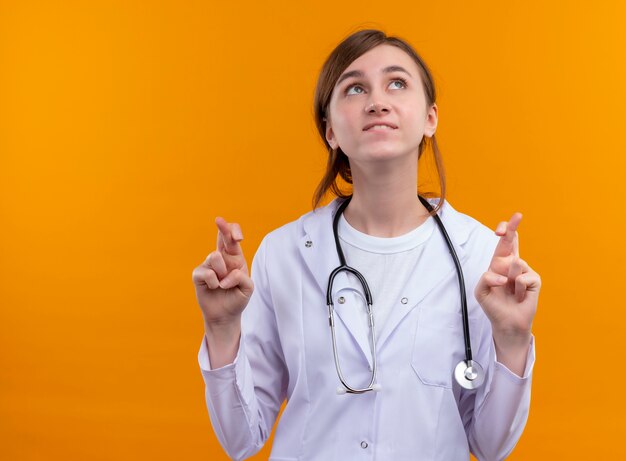 Wishing young female doctor wearing medical robe and stethoscope looking up with crossed fingers on isolated orange wall with copy space