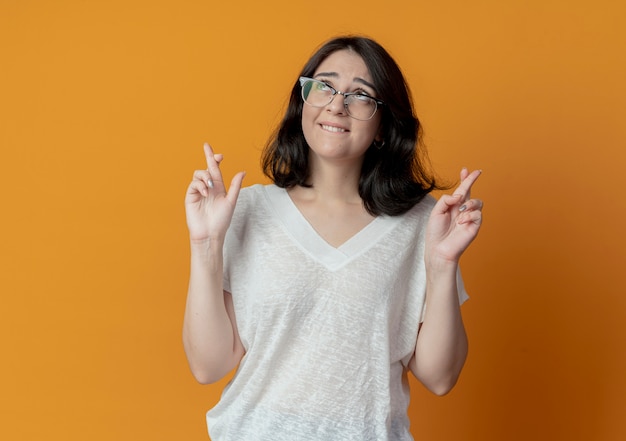 Wishing young pretty caucasian girl wearing glasses looking up and doing crossed fingers gesture isolated on orange background with copy space