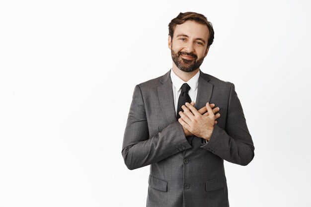 With care for our clients Smiling handsome salesman realtor holding hands on his heart and looking friendly and caring at camera standing in suit over white background