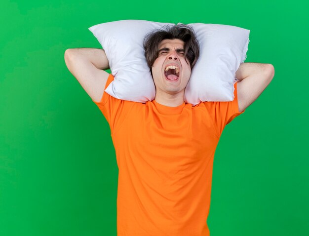 With closed eyes angry young ill man holding pillow behind head isolated on green background