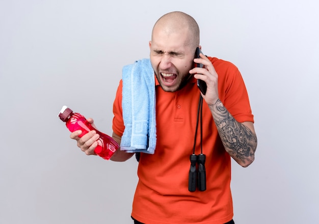 С закрытыми глазами сердитый молодой спортивный мужчина держит бутылку с водой с полотенцем на плече со скакалкой и разговаривает по телефону, изолированному на белой стене Бесплатные Фотографии