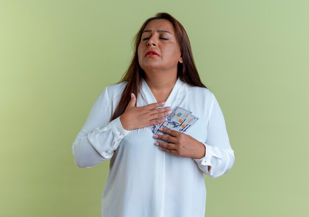 Foto gratuita con gli occhi chiusi donna di mezza età caucasica casuale che tiene contanti e che mette la mano sul cuore isolato sulla parete verde oliva