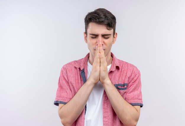 With closed eyes caucasian young guy wearing pink shirt showing pray gesture on isolated white