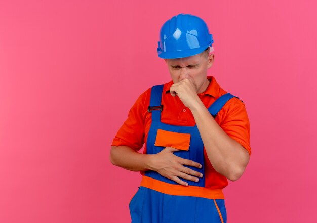 With closed eyes caughing young male builder wearing uniform and safety helmet on pink