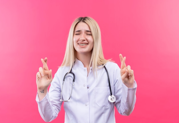 С закрытыми глазами обеспокоенная доктор молодая блондинка со стетоскопом в медицинском халате и зубной скобе скрещивает пальцы на изолированной розовой стене