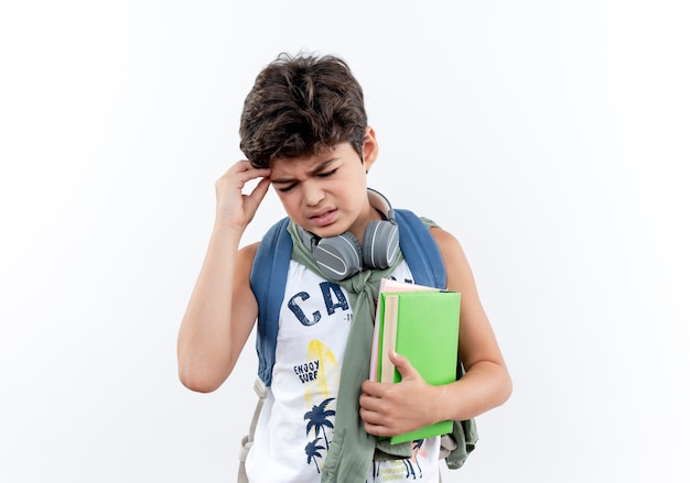 With closed eyes concerned little schoolboy wearing back bag and headphones holding book and putting hand on head isolated on white