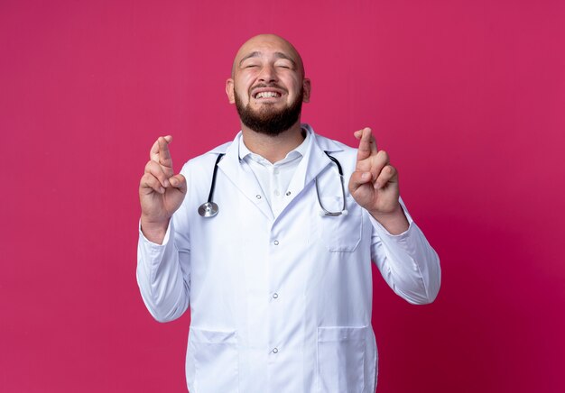 С закрытыми глазами обеспокоенный молодой лысый врач-мужчина в медицинском халате и стетоскопе скрещивает пальцы, изолированные на розовом