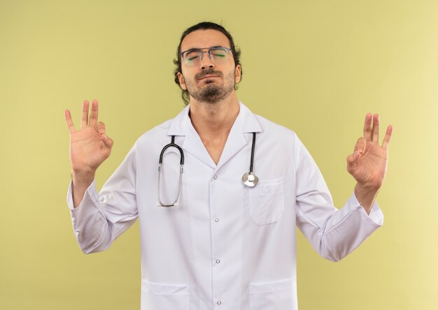 With closed eyes concerned young male doctor with optical glasses wearing white robe with stethoscope showing okey gesture on green