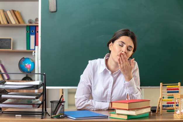 目を閉じて口を手で覆った若い女教師が教室で学校の道具を持ってテーブルに座っている