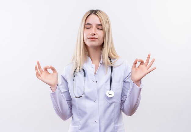 С закрытыми глазами доктор молодая блондинка со стетоскопом и медицинским халатом, показывающая хороший жест обеими руками на изолированной белой стене