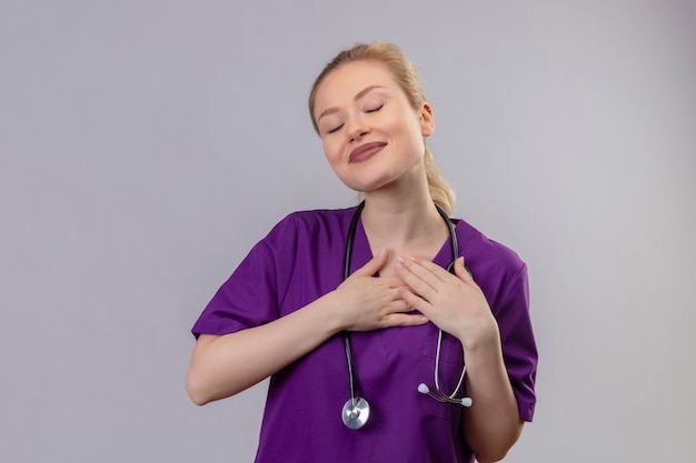 Молодая девушка с закрытыми глазами в фиолетовом медицинском халате и стетоскопе положила руки на себя на изолированном белом фоне