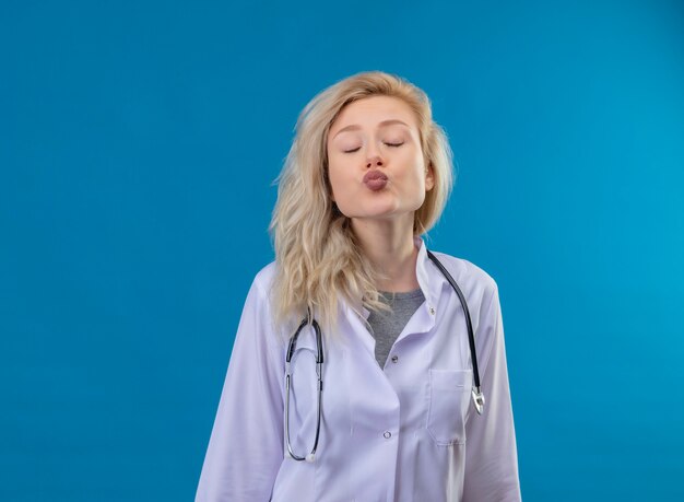 닫힌 된 눈으로 의사 어린 소녀 파란색 배경에 키스 제스처를 보여주는 의료 가운에 청진기를 착용