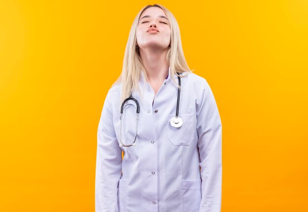 With closed eyes doctor young girl wearing stethoscope in medical gown showing kiss gesture on isolated yellow wall