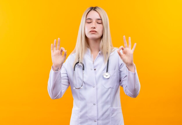 With closed eyes doctor young girl wearing stethoscope in medical gown showing okey gesture with both hands on isolated yellow wall