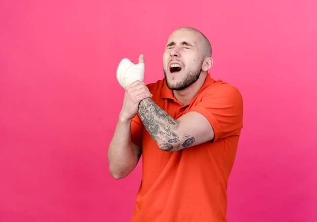 Бесплатное фото С закрытыми глазами травмированный молодой спортивный мужчина с повязкой на запястье схватил руку, изолированную на розовой стене