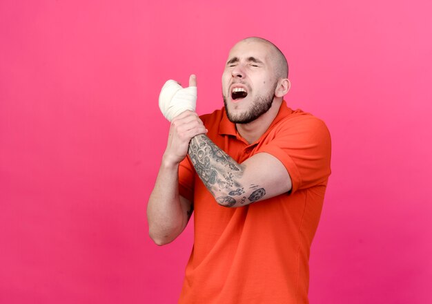 With closed eyes injured young sporty man with wrist bandage grabbed hand isolated on pink wall