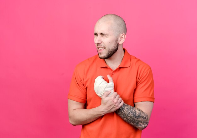 With closed eyes injured young sporty man with wrist bandage grabbed hand isolated on pink wall