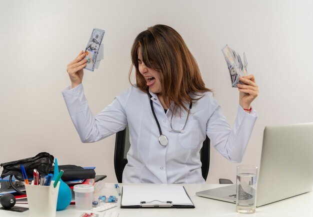 With closed eyes joyful middle-aged female doctor wearing wearing medical robe with stethoscope sitting at desk work on laptop with medical tools holding cash on white wall