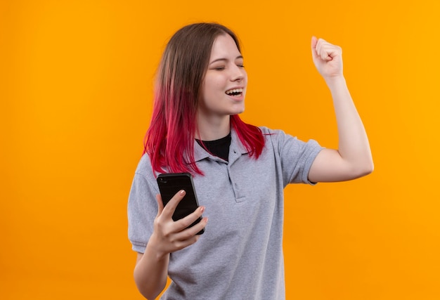 With closed eyes joyful young beautiful girl wearing gray t-shirt holding phone showing yes gesture on isolated yellow wall