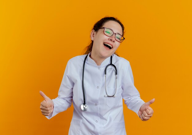 С закрытыми глазами радостная молодая женщина-врач в медицинском халате и стетоскопе в очках показывает палец вверх изолированные