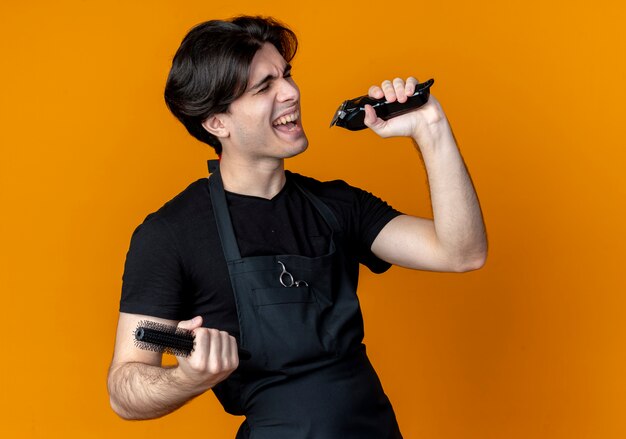 With closed eyes joyful young handsome male barber in uniform holding hair clippers with comb and singing isolated on orange