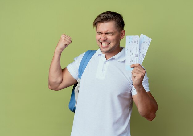 With closed eyes joyful young handsome male student wearing back bag holding tickets and showing yes gesture isolated on olive green