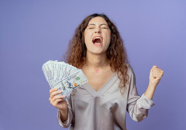 With closed eyes joyful young pretty girl holding cash and showing yes gesture isolated on blue wall