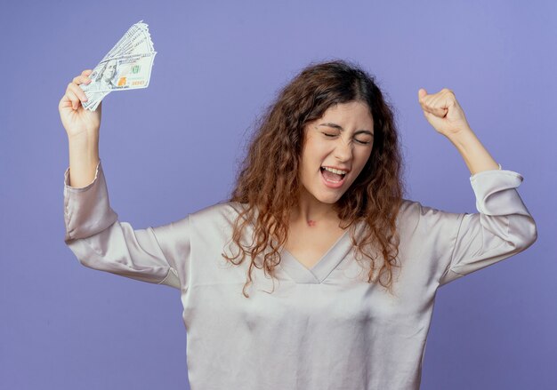 With closed eyes joyful young pretty girl raising hand and cash isolated on blue wall