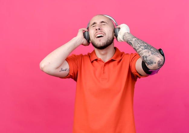 With closed eyes joyful young sporty man with wrist bandage wearing headphones with phone arm band and listen music isolated on pink wall