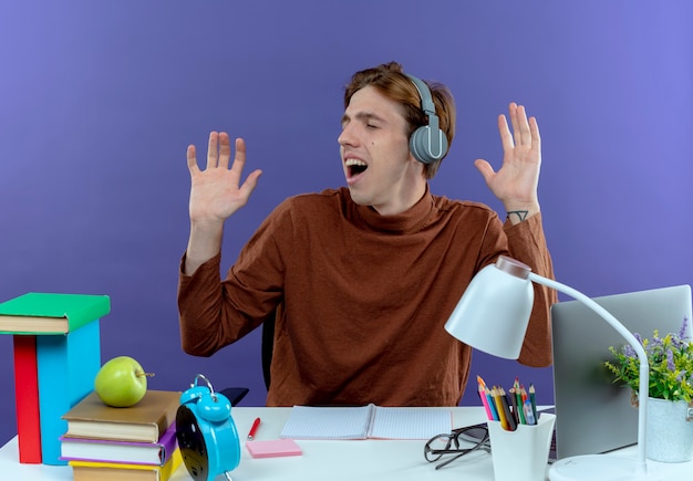 Free photo with closed eyes joyful young studend boy sitting at desk with school tools listen music on headphones and spreads hands on purple