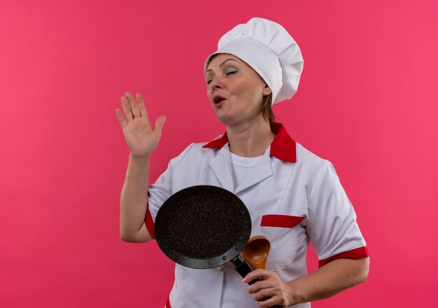 With closed eyes middle-aged female cook in chef uniform holding frying pan and sings