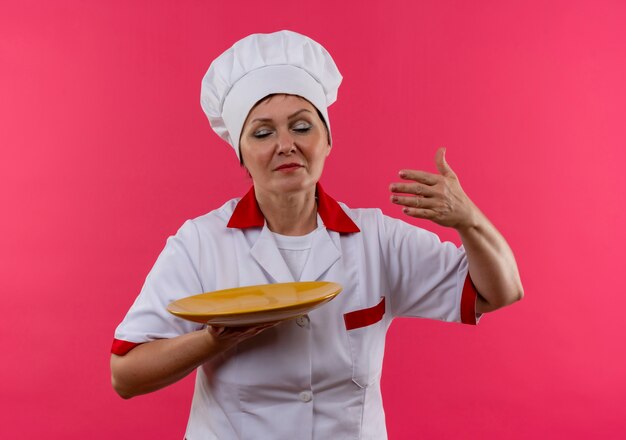 With closed eyes middle-aged female cook in chef uniform prtend sniffing holding plate