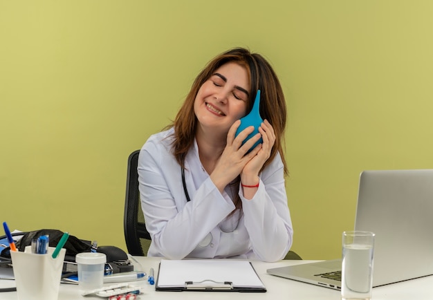 Free photo with closed eyes pleased middle-aged female doctor wearing medical robe with stethoscope sitting at desk work on laptop with nedical tools holding enema  with copy space