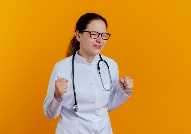 With closed eyes pleased young female doctor wearing medical robe and stethoscope with glasses showing yes gesture isolated