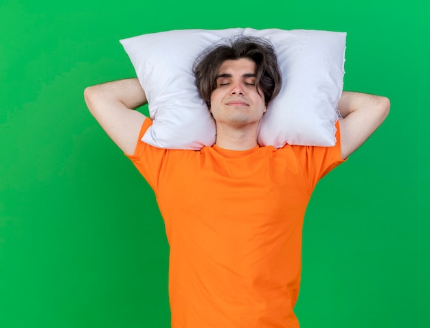 Бесплатное фото С закрытыми глазами довольный молодой больной мужчина держит подушку за головой, изолированную на зеленом фоне
