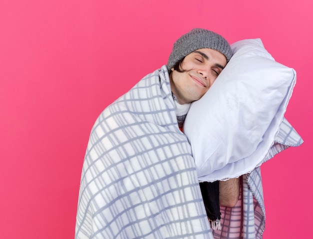 With closed eyes pleased young ill man wearing winter hat with scarf wrapped in plaid putting head on pillow isolated on pink background
