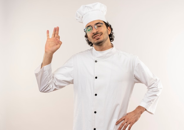 With closed eyes pleased young male cook wearing chef uniform and glasses showing okey gesture and putting hand on hip isolated on white wall