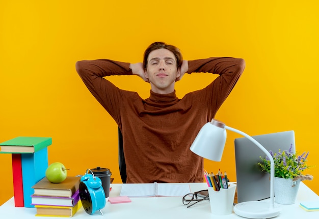 With closed eyes pleased young student boy sitting at desk with school tools keeping hand behind head on yellow