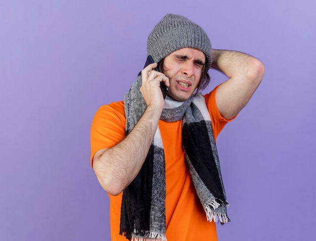 With closed eyes regretted young ill man wearing winter hat with scarf speaks on phone and putting hand behind head isolated on purple background