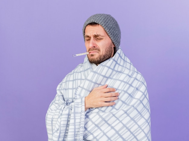 With closed eyes regretted young ill man wearing winter hat with scarf wrapped in plaid holding thermometer in mouth isolated on purple background