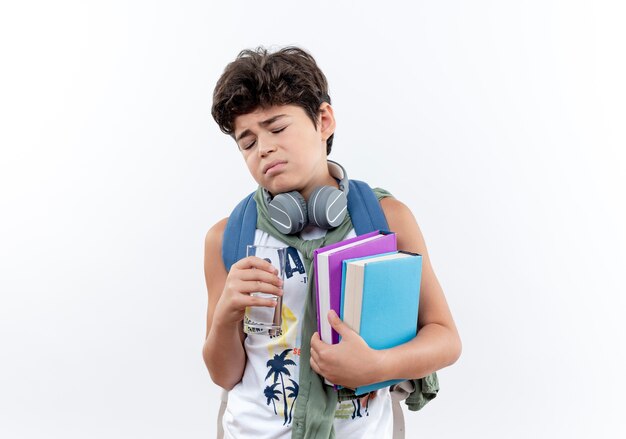 С закрытыми глазами грустный маленький школьник в задней сумке и наушниках держит стакан воды и книгу, изолированные на белом