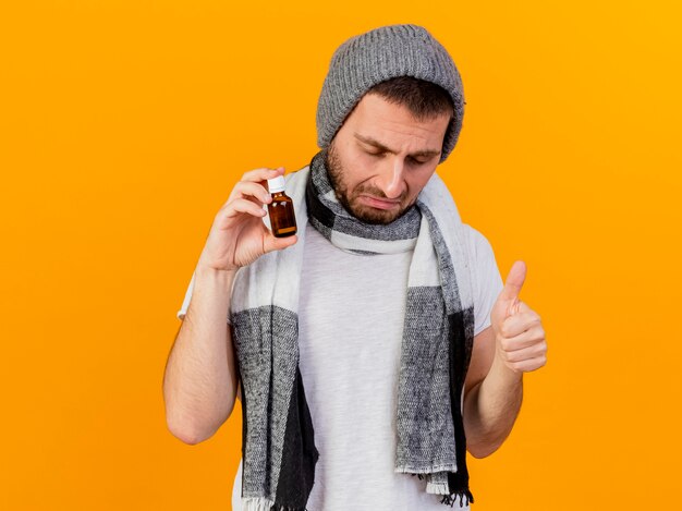 With closed eyes sad young ill man wearing winter hat and scarf holding medicine in glass bottle showing thumb up isolated on yellow background
