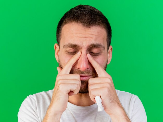 With closed eyes sad young ill man wiping eyes with fingers isolated on green background