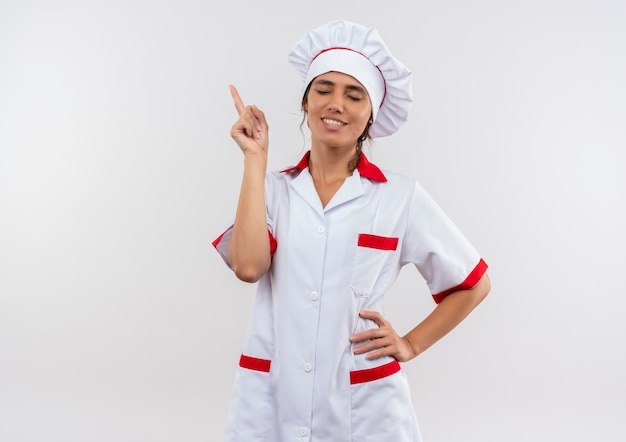 With closed eyes smiling young female cook wearing chef uniform points to up with copy space