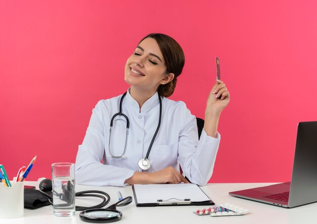 С закрытыми глазами улыбается молодая женщина-врач в медицинском халате со стетоскопом, сидя за столом, работает на компьютере с медицинскими инструментами, держа кредитную карту с копией пространства