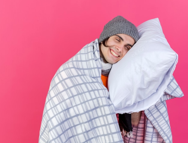 With closed eyes smiling young ill man wearing winter hat with scarf wrapped in plaid putting head on pillow isolated on pink background