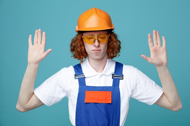 С закрытыми глазами, разводящими руками, молодой строитель в форме, изолированный на синем фоне